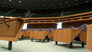 Een foto van de tweede kamer. Blauwe stoelen met een grijs logo staan achter lichtbruine tafels met zwarte poten, steeds twee stoelen per tafel. Links staat een tafel met een microfoon. Op een verhoging achteraan staan stoelen voor publiek.
