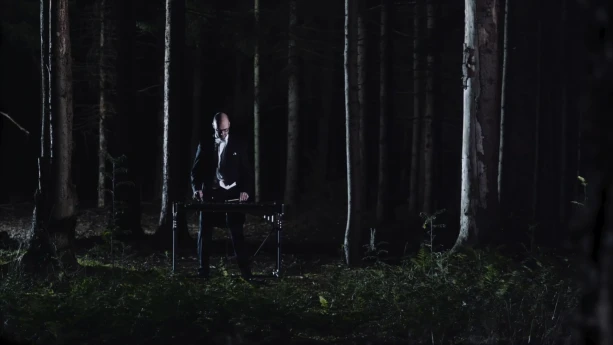 Een foto van een man in een zwart pak die in een donker bos tussen de bomen staat. Voor de man staat een instrument, een xylofoon. Dat is een soort piano met toetsen waar je op moet slaan. De man speelt op de xylofoon met houten stokken.