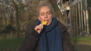 Een foto van Janouk die met haar ogen dicht buiten staat en een ijsje in haar mond steekt. Het is een waterijsje van het type rakket, waarvan de gele en oranje delen te zien zijn. Janouk draagt een donkergrijze jas en een blauwgrijze sjaal.