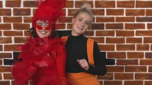 Een foto van een muur met daarvoor Janouk in oranje broek met banden om haar schouders en een zwart shirt. Ernaast staat een vrouw in een rood pak met rode veren en vormen van vlammen erop, een rode muts van veren en een zonnebril met rode glazen op. 