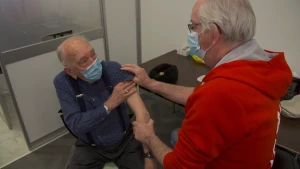 Een foto van twee mannen met een blauw mondkapje op, één man die zit en zijn mouw opstroopt en één man die zijn hand om de arm van de man heeft gelegd en zijn andere hand op de schouder van de man. Ernaast staat een tafel. De man gaat gevaccineerd worden.