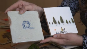 Een foto van handen met twee vierkante tegeltjes. Op één oud wit  en beschadigd tegeltje, staan in blauw twee jongetjes met vishengels. Op een wit tegeltje staan in kleur een fietsende moeder met een karretje eraan met twee kinderen erin en bomen erboven.