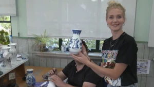 Een foto van Janouk die een Delfts blauw vaasje voor de camera houdt: wit met een blauw geschilderde opdruk en in het midden een blauw klokhuis. Erachter is iemand bezig een ander vaasje te boetseren op een tafel. Op de tafel staan ook andere vaasjes.