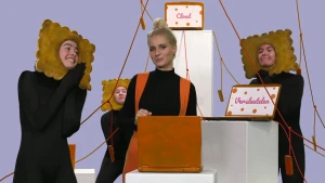 Een foto van Janouk en drie andere mensen die een zwart pak dragen en een bruin koekje met golvende randen om hun hoofd. Er hangen rode touwen waar koekjes aanzitten en er staan laptops waar vellen met de woorden "Versleutelen" en "Cloud" op geplakt zijn.