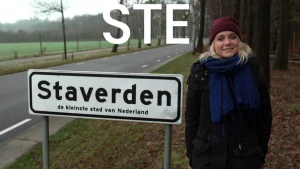 Een foto van Janouk die naast een wit bord met zwarte letters staat. Op het bord staat "Staverden, de kleinste stad van Nederland". Erboven staan de letters "S", "T' en "E" vóór de foto. Links is een autoweg te zien en achter Janouk staan bomen.