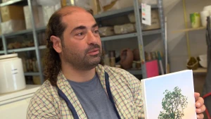 Een foto van Navid Nuur die in zijn handen een foto heeft van een boom met groene bladeren voor een blauwe lucht die middenin rood is door zonnestralen. Erachter staan rekken vol met ingepakte artikelen die hij gebruikt om zijn kunst te maken.