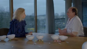 Een foto van Janouk en een dokter achter een tafel met daarop witte, door midden gedeelde bollen die op elkaar passen. Voor één paar bollen staat één groen poppetje, voor het andere paar drie groene poppetjes en voor het laatste paar drie roze poppetjes.