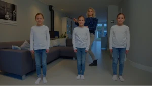 Een foto van drie meisjes die er precies hetzelfde uitzien en naar de camera kijken. Het is een eeneiige drieling. Ze dragen allemaal een witte trui, een blauwe spijkerbroek en witte sokken. Janouk staat achter het middelste meisje in de woonkamer.