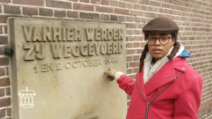 Een foto van Eva die voor een muur staat en naar een rechthoekig monument op de muur wijst. Op het monument staat "Vanhier werden zij weggevoerd - 1 en 2 october 1944". Het is een monument voor alle slachtoffers van de nazi's bij een razzia in Putten.