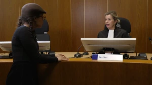 Een foto van Eva die vóór een grote tafel staat. Achter de tafel zit een rechter op een zwarte stoel in een zwart, ruim pak met een rechthoekige witte doek om de nek. De rechter zit achter microfoons, een beeldscherm en een bordje met "Rechter" erop.