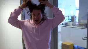 Een foto van Pascal die in de camera kijkt, twee plukken van zijn haar vastheeft en deze omhoog houdt boven zijn hoofd.  Hij staat voor een deur van glas, waarachter een laboratorium te zien is met bakjes en een wit rechthoekig ding op een witte kast.  