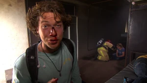 Een foto van Pascal die een nagemaakte wond met rood bloed op zijn neus heeft zitten. Het bloed loopt van bovenaan zijn neus naar zijn neusgaten. Erachter zit een vrouw op de grond die acteert dat ze gewond is naast een brandweerman in brandweerkleding.