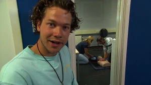 Een foto van Pascal die voor een kamer staat met daarin een op de grond zittende man en vrouw naast een plastic menselijk bovenlichaam, van hoofd tot buik zichtbaar. De man drukt op de borst van de pop, de vrouw opent een klepje van een apparaat.