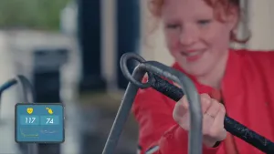 Een foto van een meisje met een zwarte stok met een ronde, stalen cirkel erop. Ze beweegt het gedeelte met de stalen cirkel om een stalen stang met bochten, een spiraal heen. Linksonder staat een getekend hart met waarde 117 en een zweetdruppel met 74.