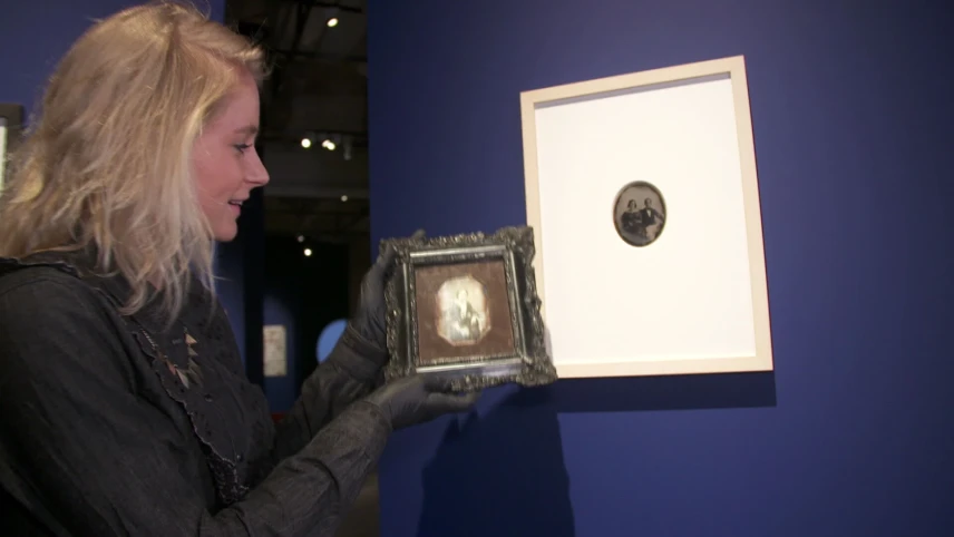 Een foto van presentator Janouk. Ze staat in een museum en houdt een hele oude foto vast, die in een zilveren lijst zit. Aan de muur hangt ook een hele oude zwart-witfoto. 