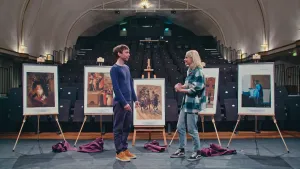 Een foto van Janouk en Fabian op een podium met vijf schildersezels of schilderij standaarden met vier kopieën van schilderijen en één foto. De tekst eronder is niet leesbaar. Te zien zijn: katten, mensen, mannen te paard, een gebouw en een vrouw.