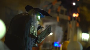 Een foto van een pop van een heks met grijs haar. De huid van gezicht en handen is groen. Het zichtbare oog met rode pupil licht op in het donker. Ze draagt een zwarte cape, een zwarte puntmuts en in haar handen een boek, waarschijnlijk een toverboek.