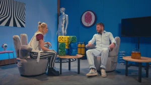 Een foto van Janouk en prof op een grijze stoel in de vorm van een hand met twee vingers als armleuning en twee vingers als rugleuning. Tussen hen in staat een kleine, ronde tafel met gele, groene en blauwe dobbelstenen, van iedere kleur drie.