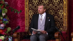 Een foto van koning Willem-Alexander in een donker pak met lichtpaars vest eronder. Hij zit op een grote, houten troon met op de rug goudkleurige bloemvormen op een zwarte achtergrond. Er staan microfoons voor en de koning heeft een stapel papieren vast. 