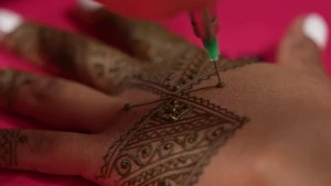 Een foto van een hand met bovenop een donkerbruine versiering, een tijdelijke henna tatoeage. Het zijn rechte en gekrulde lijnen in een vierkante vorm, lijnen die ook op de vingers zitten. Er prikt een naald in de hand bij het zetten van een nieuwe lijn.