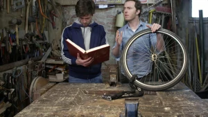 Een foto van de presentatoren Pieter en Willem in een werkplaats. Willem kijkt in een groot boek. Pieter heeft een voorwiel van een fiets vast. Dat staat op een tafel. Aan het wiel zit een groot kettingslot.