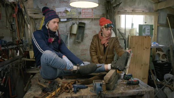 Een foto van presentatoren Willem en Pieter die in een schuur zijn. Willem zit op tafel met een paar schaatsen aan.
