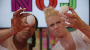 Een foto van Eva en Janouk die allebei een wit ei voor de camera houden. Eva kijkt met een normale blik naar het ei, Janouk heeft haar mondhoeken iets omlaag. Ze kijken of aan de buitenkant zien of het ei rauw of gekookt is.