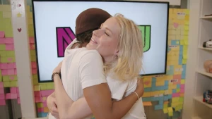 Een foto van Eva en Janouk die elkaar omhelzen. Janouk's gezicht is te zien en ze heeft een lach op haar gezicht. Erachter hangt een beeldscherm en op de muur hangen allemaal vierkante, gekleurde briefjes die post-its genoemd worden.