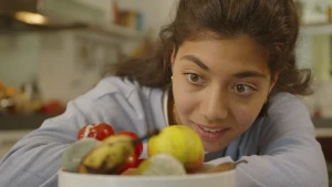 Een foto van een presentator die kijkt naar fruit in een witte schaal: appels, een banaan, tomaten en een sinaasappel. Op twee stuks fruit zijn groenwitte vlekken te zien. Dit is schimmel, dat komt op eten te zitten dat bedorven en niet meer eetbaar is. 