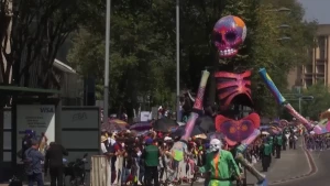 Een foto van een optocht bij de viering van de Dag van de Doden in Mexico. Er is een grote, kleurrijke pop van een skelet te zien en iemand die ervoor loopt met groene en oranje kleding en een grijs doodshoofd masker. Mensen achter hekken kijken ernaar.