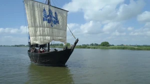 Een foto van een schip van zwartgekleurd hout in het water. Het schip heeft één groot wit zeil, waarop in een blauwe kleur een kasteel staat met drie torens. Er zijn van veraf drie zeemannen op het schip te zien in rode jas en met een zwarte hoed. 