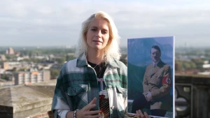 Een foto van Janouk die voor een stenen muur op een dak van een gebouw staat. Ze heeft in haar linkerhand een foto van nazi leider Adolf Hitler, zittend op de rand van een balkon in een berggebied, met lichtbruine jas en een rode nazi band om zijn arm. 