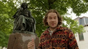 Een foto van Pascal die buiten staat met bomen en een gebouw op de achtergrond. Pascal wijst met zijn duim naar een beeld van Spinoza. Spinoza zit, heeft zijn vuist onder zijn kin en denkt na. Hij was een bekende denker, een filosoof in de 17e eeuw.