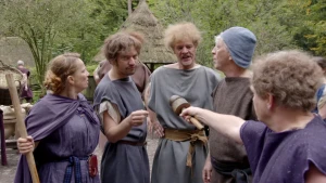 Een foto van een interviewer met een soort microfoon en vier andere mensen: een vrouw in een blauw kostuum met een stok in handen, een man in een grijs kostuum, een man in een lichtgrijs kostuum en  een man met een bruin kostuum en een blauwgrijze hoed.