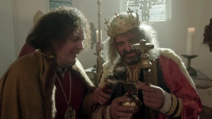 Een foto van een man die een andere man interviewt met een zilverkleurige microfoon. De ander is verkleed als Karel de grote in een zwart met rood vest, cape met witte bondranden, een kroon met edelstenen en hij heeft twee dingen vast met een kruis erop.