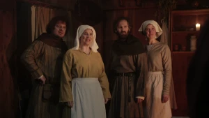 Een foto van vier mensen die naar iets rechtsachter de camera kijken. De twee mannen dragen een bruin kostuum met zwarte rand om hun nek en schouders. De twee vrouwen dragen een lichtbruine trui, blauwe en lichtbruine rok en een witte doek om hun hoofd.