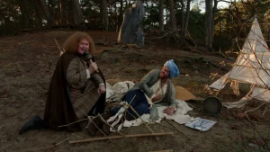 Een foto van een interviewer en een man verkleed als Ibn Firnas die op een witte doek ligt met een blauwe doek, een tulband, om zijn hoofd en zijn rechterhand op zijn buik. Een rek van stokkken ligt op zijn benen en zit ook aan een tent erachter vast. 