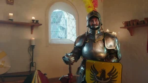 Een foto van een man in een metalen harnas met een metalen helm op, waarvan de klep openstaat. Op de helm zit een gele en een rode veer. De man heeft in zijn ene hand een zwaard en in zijn andere een geel schild met een zwarte vogel met een kroon erop.
