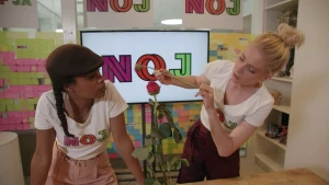 Een foto van Eva en Janouk die achter een tafel staan. Op de tafel staat een bloem rechtop, een rode roos. Janouk kijkt naar de bloem en heeft zelf een andere rode roos vast aan de groene steel. Eva en Janouk dragen een t-shirt met "NOJ" erop.