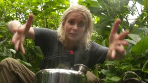 Een foto van Janouk die achter een stalen fluitketel zit in een kas met groene planten. Een fluitketel is een ketel om water mee te koken en als het kookt, komt uit de ronde opening een fluitend geluid. Janouk heeft haar handen wijd uit elkaar.