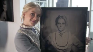 Een foto van Janouk die lacht naar de camera en een zwart-wit foto van zichzelf in handen heeft, die met een hele oude camera is gemaakt. Ze heeft een grijze jurk aan en om haar nek een witte lap stof met bloemetjes erop met een blauwe strik erom.