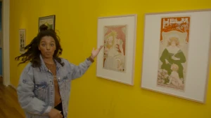 Een foto van Sosha die met haar hand wijst naar twee witte schilderijlijsten op een gele muur. In de lijsten zitten tekeningen: één van een vrouw die een kind een lepel cacao of chocolade van het merk Helm geeft, één van een naar beneden kijkende vrouw.