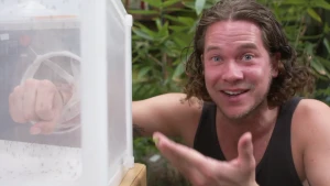 Een foto van Pascal in een zwart hemd. Hij heeft zijn rechtervuist door een rond gat gestoken in een vierkante doos met wit insectengaas aan de zijkanten. De doos zit vol met kleine insecten, muggen, die mensen en dieren steken en hun bloed opzuigen.