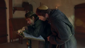 Een foto van twee mannen in een pei een zwart pak met capuchon.  Het zijn monniken, die samen wonen en geloven in god. De ene man staat gebogen over een tafel en schrijft in een boek. De andere man kijkt over zijn schouder mee wat hij opschrijft.