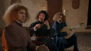 Een foto van drie mannen, minnezangers, in middeleeuwse kleding, bruin, zwart en blauw van kleur. De voorste man heeft een trommel vast, de middelste man een ronde, bruine fluit en de rechterman een lichtbruine luit, een middeleeuwse gitaar. 