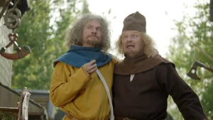 Een foto van twee mannen. De linkerman in een bruingeel kostuum  met een blauwgrijze doek om de nek heeft een staafje in zijn hand en zijn andere hand om de nek van de rechterman. Die draagt een bruin kostuum en hoed en kijkt scheel en bang omhoog. 