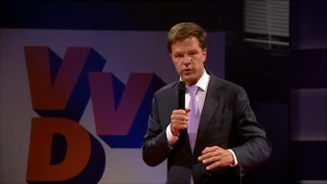 Een foto van Mark Rutte, minister-president van Nederland. Op de achtergrond hangt het oranje met blauwe logo van de politieke partij VVD. Mark heeft een zwarte microfoon in zijn rechterhand, draagt een zwart pak, witte blouse en een lichtpaarse stropdas.