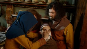 Een foto van twee mannen die toekijken hoe een jongen in een bruin kostuum, waarvan het gezicht niet te zien is door een donkerblauwe kap, een man op een stoel aan het scheren is met een mes. De man op de stoel heeft wit scheerschuim op zijn gezicht. 