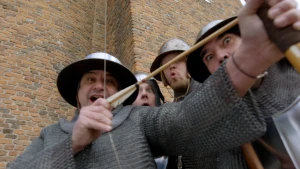 Een foto van vier soldaten die een maliënkolder, een beschermend vest van metalen ringetjes, aanhebben en een metalen helm op. De voorste soldaat lacht naar de camera en trekt aan het witte touw van een houten boog met een pijl met witte staartveren erop.