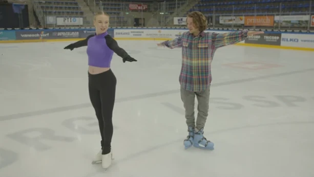 Een foto van de zestienjarige kunstschaatser Lindsay van Zundert. Ze staat samen met de presentator binnen in een hal op het ijs. Ze hebben schaatsen aan en staan in dezelfde houding, met hun armen gestrekt opzij.